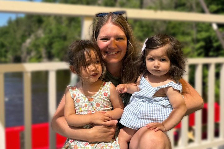 Woman smiling with two young girls on a sunny day, outdoors with trees in the background.