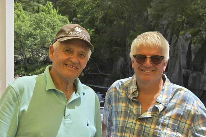 Two men smiling on a boat with a river and forested cliff behind them.