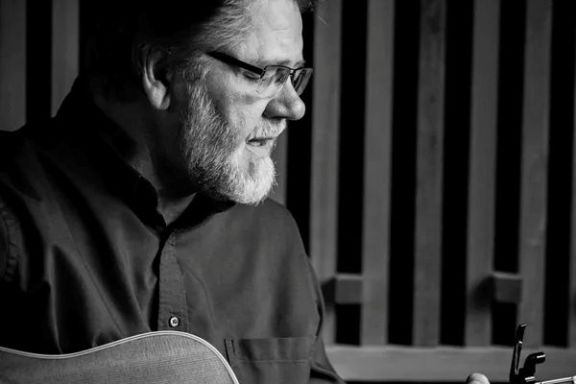 Man with glasses playing an acoustic guitar in a dimly lit room.