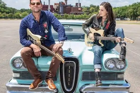 Two people with guitars sitting on a vintage car, industrial backdrop.