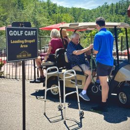 Three people on a golf cart at a scenic loading area with a walker nearby.