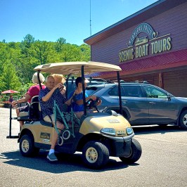 People on a golf cart near a scenic boat tours building under a clear blue sky.