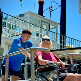 Man assists woman in wheelchair on a boat ramp under clear sky.