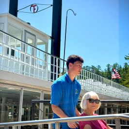 Young man pushing an older woman in a wheelchair on a boat deck with an American flag and clear sky.