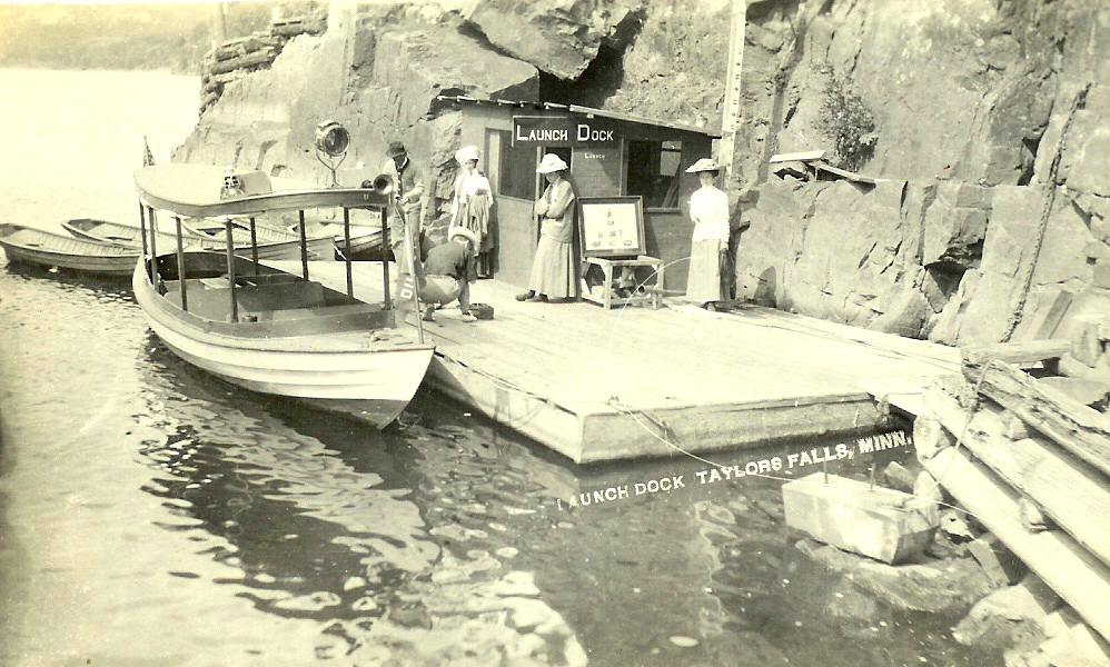People on a wooden dock beside a small boat at Taylors Falls, MN, with rocky cliffs in the background.