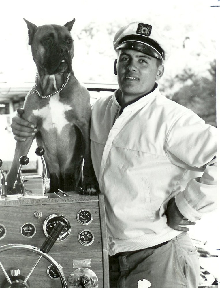 Man in a captain's hat and dog by a boat's controls, in black and white.