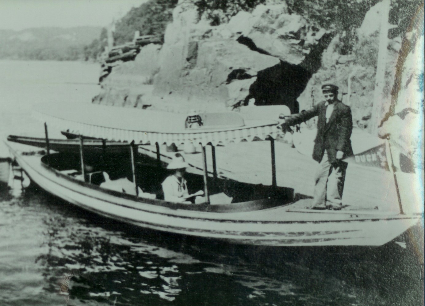 Vintage boat with two men, one standing on deck, near rocky shore.