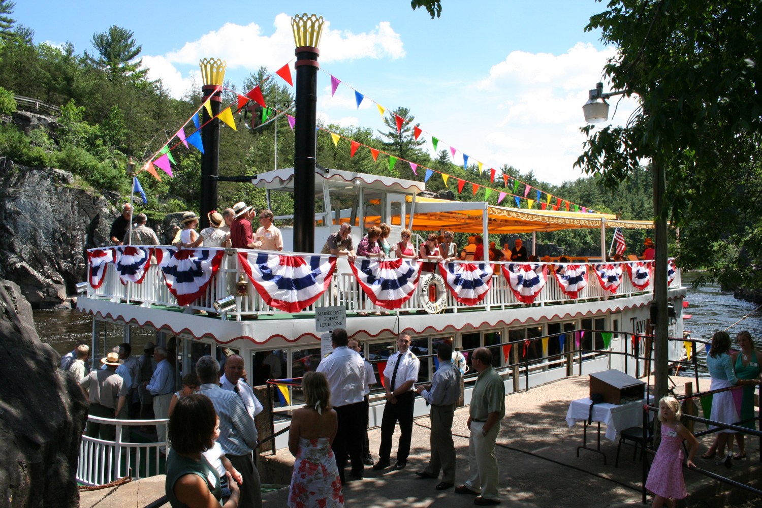 People in festive attire boarding a decorated riverboat with colorful flags and bunting in a wooded area.