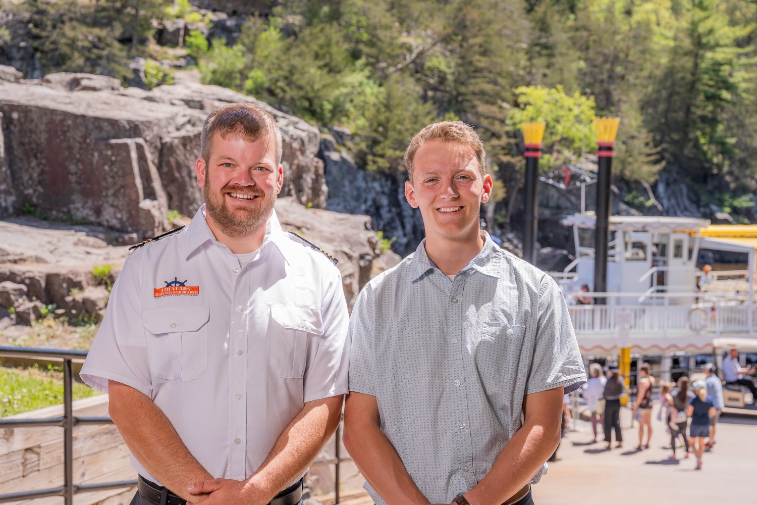 Two men smiling outdoors with a riverboat and greenery in the background.