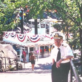 Captain in uniform stands near a path by a decorated riverboat.