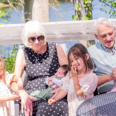 Elderly couple sitting with four young children by a wooden fence.