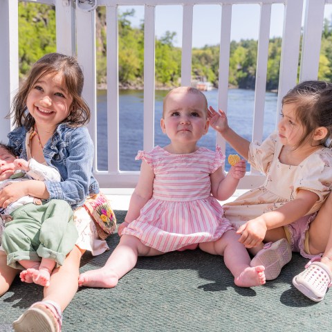 Four children sitting on a porch, three smiling, with a scenic river view in the background.