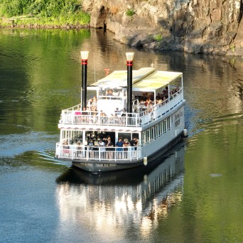 Paddle steamer with passengers sailing on a river near a rocky cliff.