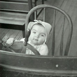 Baby in a bonnet, sitting on a chair, holding a ship's wheel playfully.
