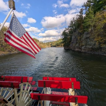 Paddle boat on a river with an American flag, surrounded by forested cliffs under a blue sky with clouds.