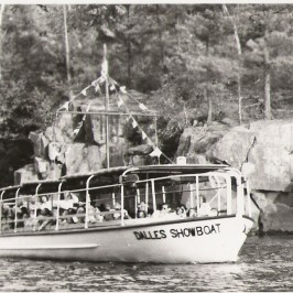 Vintage boat named 'Dalles Showboat' with passengers, near rocky shoreline and trees.