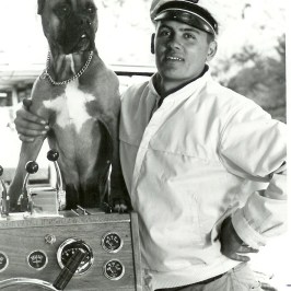 Man with a dog on a boat, both looking ahead, man in a captain's hat.