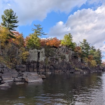 Rocky cliff with autumn trees by a calm river under a blue sky.