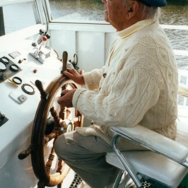 Elderly man with cap steering a boat on a river.
