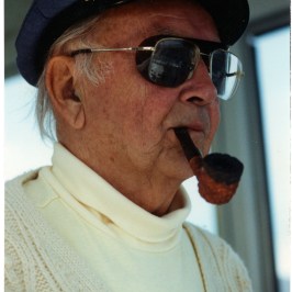 Elderly man in captain's hat and sunglasses, smoking a pipe.