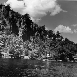 Steamboat near rocky cliff with trees and cloudy sky.