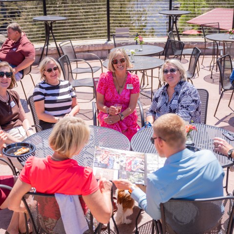 Group of people sitting at outdoor tables, talking and reading newspaper.