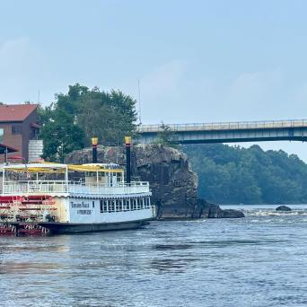 Riverboat docked near a bridge on a calm river with trees in the background.