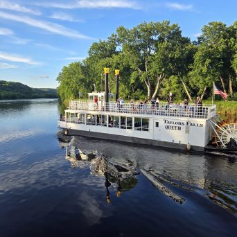 Riverboat with passengers on calm water, trees in background, American flag visible.