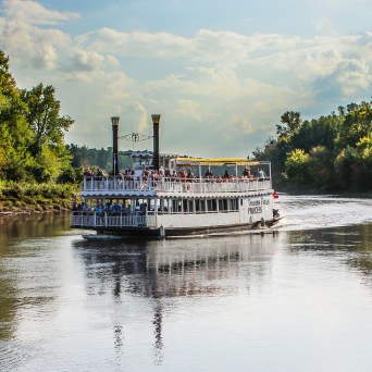 Paddle steamboat cruising on a river surrounded by trees.