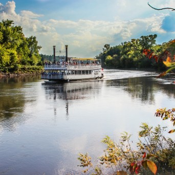 Paddleboat cruising on a tree-lined river under a blue sky with fluffy clouds.