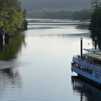 Riverboat sailing on a calm river with forested cliffs and trees.