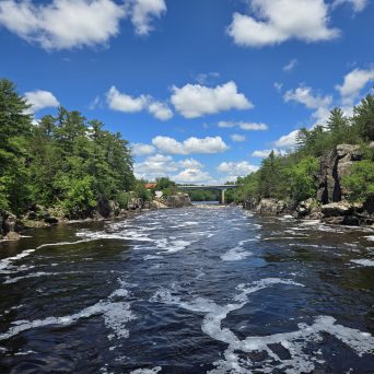River with rapids and trees under a blue sky with clouds.