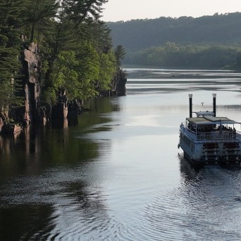 Riverboat cruising on a forested river with rocky cliffs and trees.