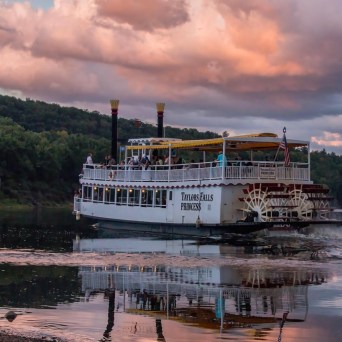 Riverboat with a paddlewheel on a calm river under a cloudy sky.