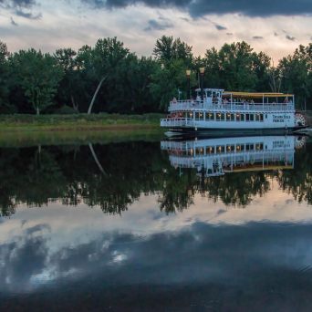 Riverboat on calm water with trees and cloudy sky in the background.