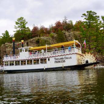 Riverboat named Taylors Falls Princess cruising on a river with rocky shore and trees in the background.