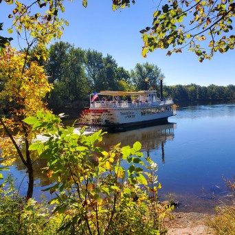 A riverboat cruises on a calm river surrounded by trees under a clear blue sky.