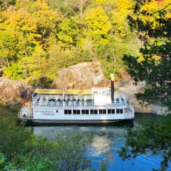 Riverboat on a wooded river with colorful autumn trees.