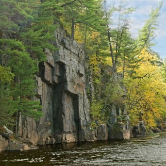 Cliffside with autumn trees by a river under a cloudy sky.