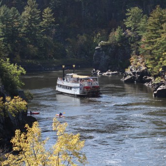 A boat travels on a river surrounded by trees and rocky cliffs, with kayakers in the foreground.
