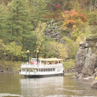 Paddle steamboat on a river surrounded by trees and rocky cliffs.
