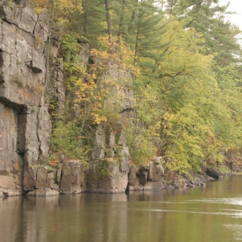 Rocky cliff beside a calm river with two kayakers, surrounded by lush green trees.