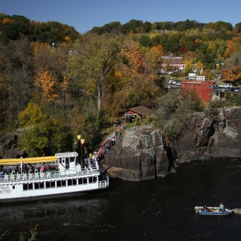 Riverboat near rocky shore with autumn trees and a small boat in the water.